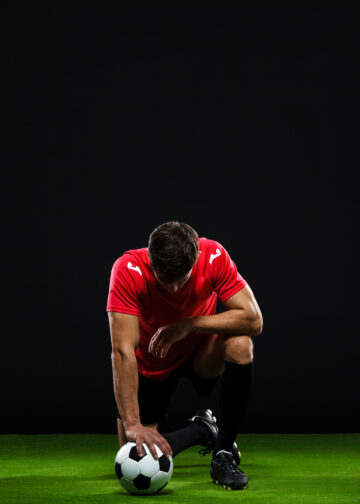 Soccer player in black and red uniform with ball standing on one knee over black background with head down